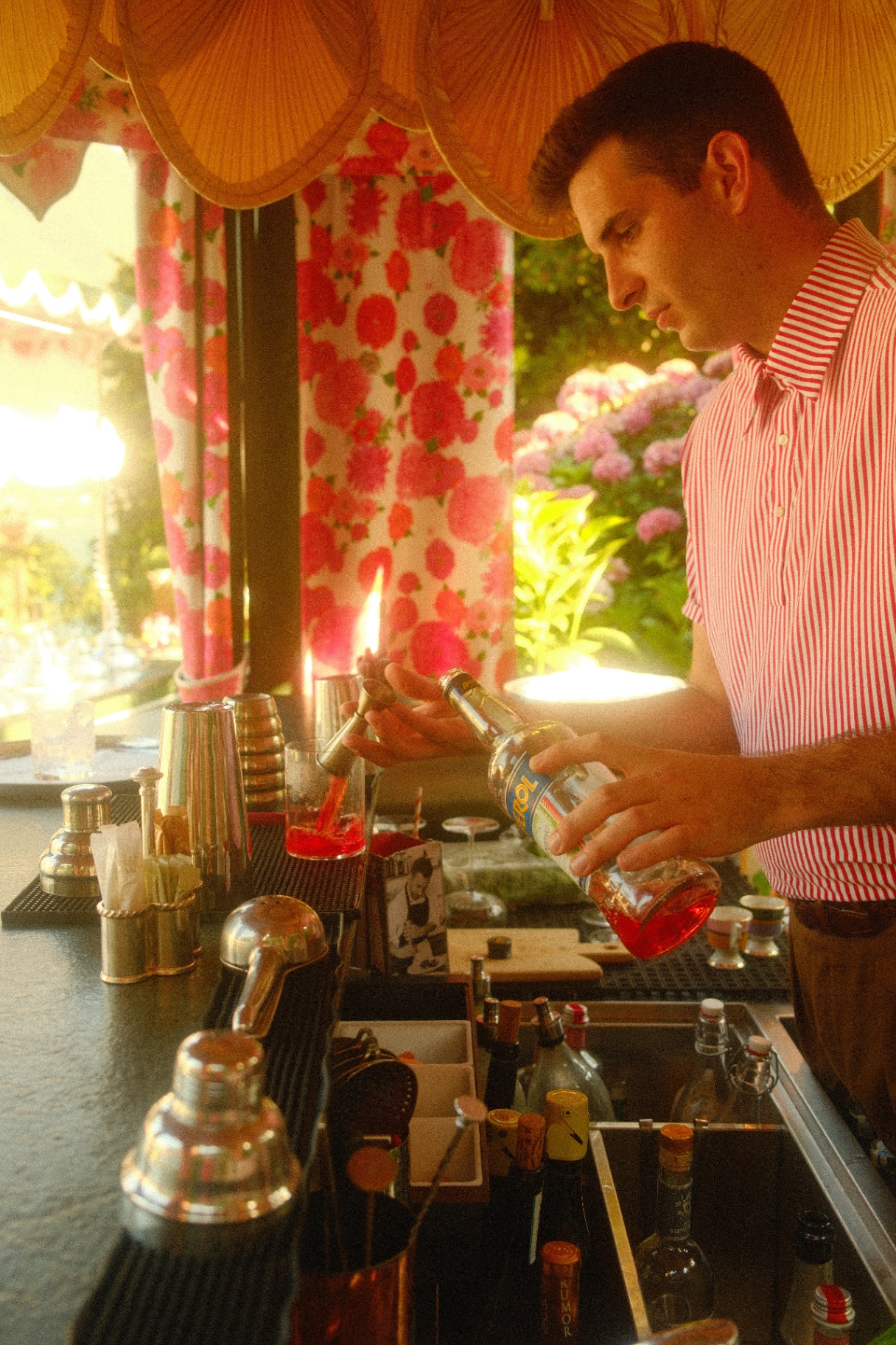 Bartender pouring an Aperol cocktail under scalloped lamps with rose-patterned curtains
