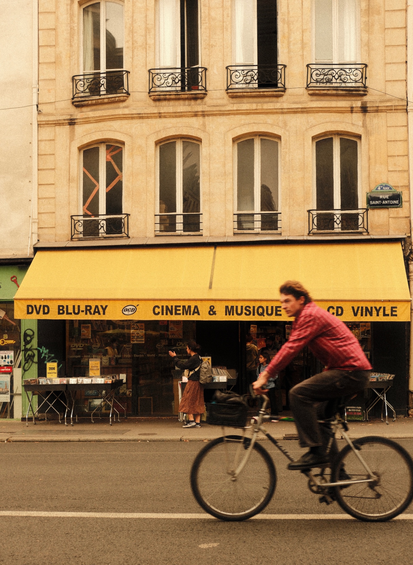 Cyclist passing a yellow cinéma & musique shopfront on rue Saint-Antoine, Paris
