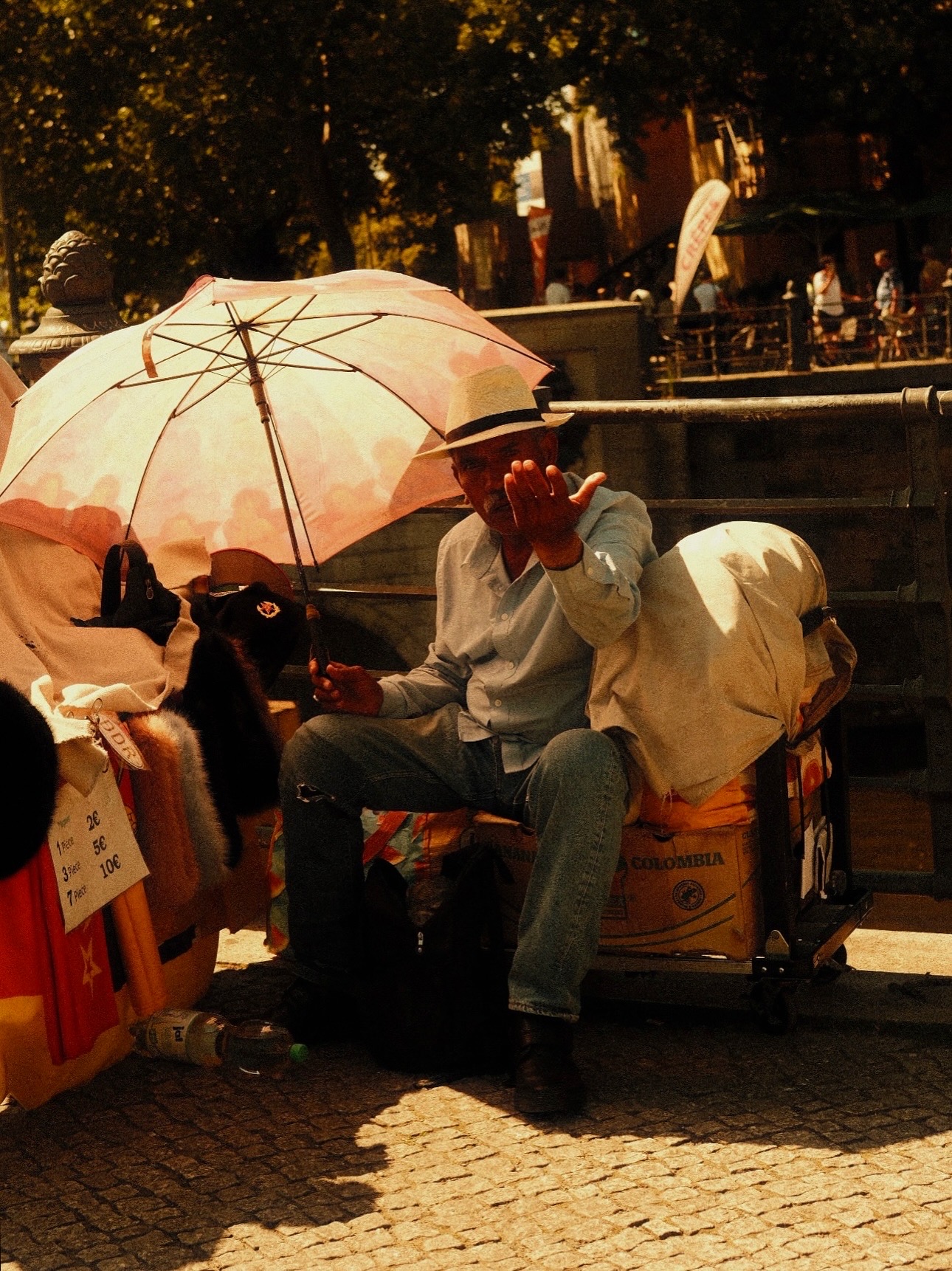 A man under a pink umbrella beside a market stall in golden light