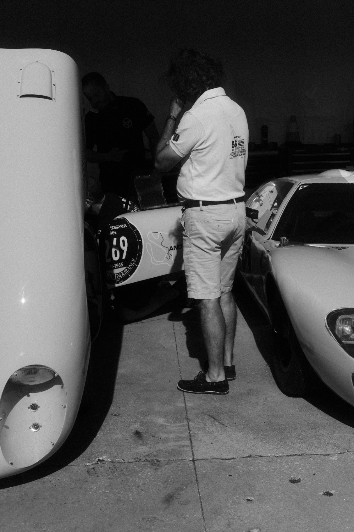 Man in shorts between two white classic race cars in the paddock