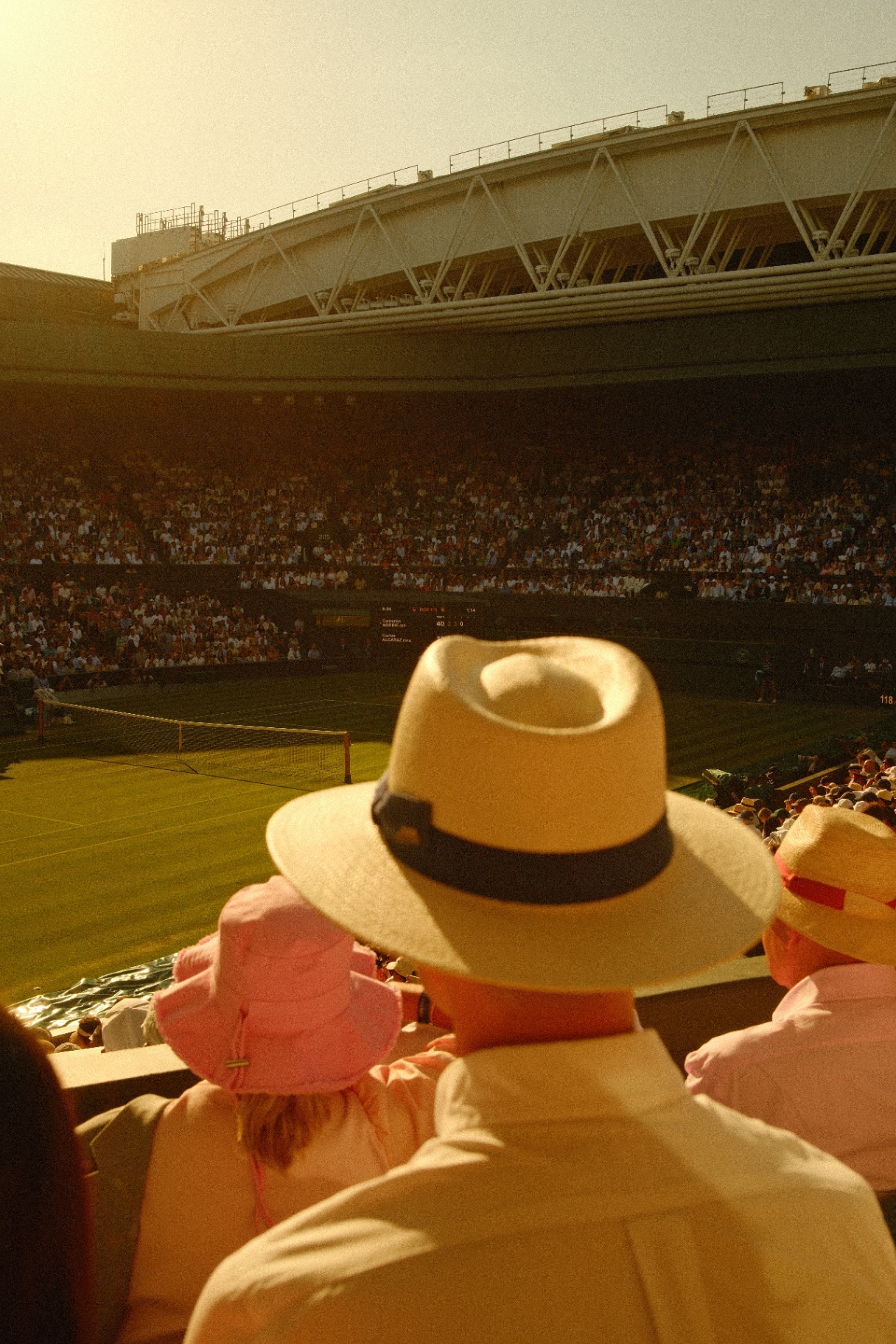 Spectators in straw hats watching a Wimbledon match in golden evening light